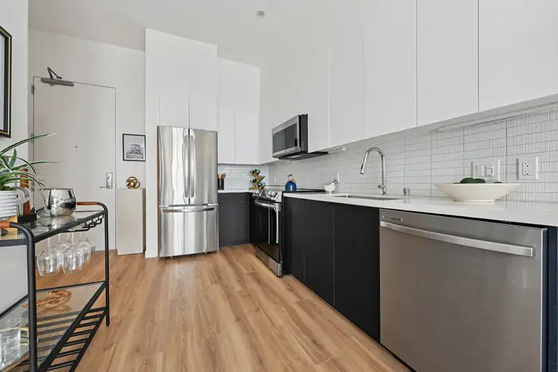 Modern Kitchen With Stainless Steel Appliances, White Tile Backsplash, Black Lower Cabinets, And Wood Flooring.